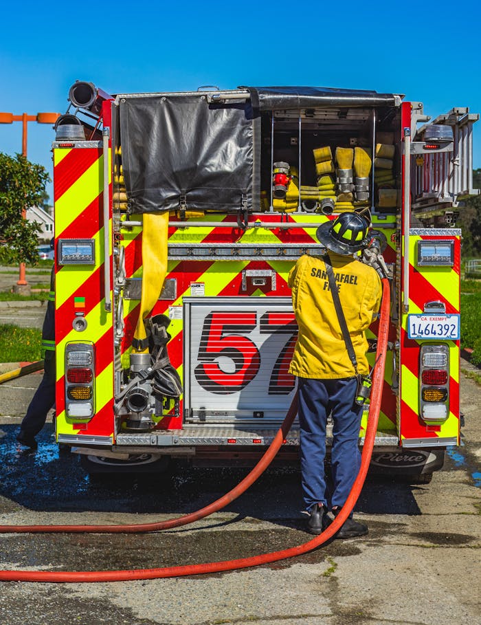 Firefighter in uniform handles hoses at the back of a fire truck outdoors.